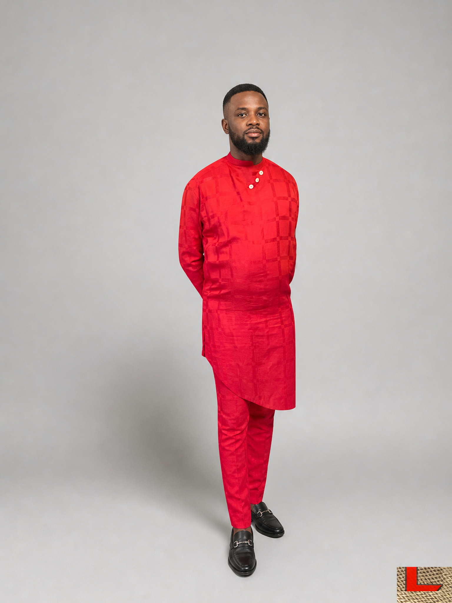 TL-branded edited portrait against a neutral grey studio background: a man wearing a long red tunic, matching red trousers and dark shoes